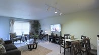 This single-floor residence offers an open-concept layout that seamlessly integrates the living, dining, and workspace areas into one cohesive environment. The living area is anchored by a large wall-mounted flat-screen television positioned above a dark wood media console, complemented by a round glass-top coffee table set atop a textured area rug over neutral carpeting. Track lighting installed on the ceiling provides versatile, directional illumination throughout the space.

A compact desk area tucked along one wall accommodates a desktop computer setup with a task lamp, making efficient use of the available space. Adjacent to it, a patterned accent chair adds a touch of visual interest near a window dressed with horizontal blinds, allowing natural light to filter in. Lush indoor tropical plants introduce organic warmth and color against the soft green and grey-toned walls.

The dining area, positioned near the living space, features dark wood chairs and a light-colored table, maintaining a consistent dark wood aesthetic throughout. The textured ceiling and overall neutral palette create a calm, functional ambiance suited for everyday living.