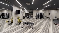 This fitness center offers a thoughtfully designed workout environment finished in a contemporary monochromatic palette of gray, white, and black, accented by bold yellow details on the free weights. The space is illuminated by striking linear LED lighting recessed into a dark ceiling, creating a polished, energetic atmosphere throughout.

The main workout area is generously proportioned, providing ample room to move between equipment zones. The cardio section features a row of treadmills and a stationary bike, each equipped with individual digital display screens. Adjacent to the cardio area, a multi-function cable machine offers versatile strength training options, while a dedicated free weight zone includes a dumbbell rack, adjustable bench, medicine balls, and ab rollers. A wall-mounted flat-screen television and curated wall art contribute to the refined aesthetic.

A glass-walled studio or stretching room is accessible through sliding glass doors, offering a bright, open area ideal for yoga, stretching, or group exercise. A water fountain is conveniently located nearby. The entire space is finished with durable striped carpet, tying together the clean, modern design throughout.
