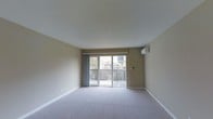 This single-floor residence offers a clean, functional layout with a welcoming and understated aesthetic. The main living area is finished with wall-to-wall carpeting in a neutral tone, providing a comfortable and cohesive look throughout. Warm beige walls complement the light-colored flooring, creating a calm and inviting atmosphere.

A standout feature of the living space is the large sliding glass door, which floods the room with natural light and provides direct access to a private outdoor deck. The deck is enclosed with a decorative lattice railing, offering a sense of privacy while maintaining an open, airy feel. Views of surrounding greenery and neighboring structures are visible through the glass, adding a pleasant outdoor connection to the interior.

The room is equipped with a wall-mounted air conditioning unit, ensuring year-round climate comfort. The smooth, white ceiling adds to the sense of openness and height within the space. Vertical drape panels frame the sliding door, offering light control and a soft finishing touch to the room.

Overall, the space presents a practical and well-lit environment with thoughtful design elements that balance comfort and simplicity, making it well-suited for everyday living.