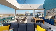 This rooftop terrace at 1725 W Katella Ave in Orange, California, offers a well-appointed outdoor living experience designed for both relaxation and entertaining. The space is open to the California sky and benefits from abundant natural sunlight throughout the day, with string lights overhead adding ambiance for evening gatherings.

The terrace is divided into several distinct zones. A fully equipped outdoor kitchen anchors one side, featuring a stainless steel built-in grill set against a striking geometric mosaic tile backsplash, complemented by bold blue cabinetry and an adjacent wooden prep and dining table with bench seating. The teal accent wall adds a vibrant pop of color to this cooking area.

A generous lounge zone offers deep, cushioned sectional seating in neutral tones, accented with colorful geometric throw pillows. Woven rattan accent chairs and small side tables create intimate conversation areas throughout the space. A high-top bar counter with teal stools lines the open perimeter railing, offering unobstructed panoramic views of the Orange County skyline and surrounding landscape.

The clean, large-format tile flooring ties the entire terrace together, while carefully placed planters with ornamental grasses and succulents add natural texture. The overall design balances modern aesthetic with a casual, social atmosphere ideal for outdoor living in Southern California.