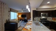 This single-floor residence offers a thoughtfully designed open-concept layout that seamlessly connects the kitchen and living areas. The kitchen is a standout feature, showcasing granite countertops that extend into a spacious island with a double undermount sink, pendant lighting with cascading cylindrical fixtures, and dark espresso cabinetry. Stainless steel appliances, including a microwave and gas range, complement the tile backsplash for a polished, contemporary finish.

The living area flows naturally from the kitchen, anchored by warm terracotta-toned accent walls that create an inviting ambiance. A ceiling fan provides both function and style overhead, while a wall-mounted flat-screen television serves as the focal point of the entertainment space. The seating arrangement includes a neutral-toned sofa and accent chairs, paired with a round glass coffee table atop a patterned area rug, lending a refined yet relaxed atmosphere. Natural light filters in through a large window dressed with valance treatments, brightening the space during daytime hours. The combination of hardwood-style flooring and soft furnishings ties the entire space together, creating a cohesive and comfortable single-floor living environment.