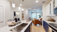 This single-floor residence showcases a thoughtfully designed open-concept layout that connects the kitchen, dining, and living areas into one cohesive space. Light wood flooring flows throughout, creating warmth and visual continuity across the main living areas.

The kitchen is a standout feature, distinguished by its two-tone cabinetry pairing bold navy lower cabinets with crisp white upper cabinets, complemented by a white hexagonal tile backsplash and light stone countertops. A central island with a stainless steel undermount sink is illuminated by stylish pendant lighting, while stainless steel appliances complete the functional design.

The living area is bathed in natural light from large windows and benefits from track lighting overhead and a ceiling fan. The space is open and inviting, with room for comfortable seating arrangements.

The bedroom is a tranquil retreat, featuring plush carpeting, a large window with blinds for natural light control, and ample wall space for décor.

The bathroom maintains the navy cabinetry theme, featuring a generous vanity with stone countertops, a large mirror, upper storage cabinets, and a glass-enclosed shower, all finished with neutral tile flooring that ties the design together cohesively.