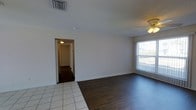 This single-story space presents an open living area that transitions seamlessly between two distinct flooring materials — classic light-toned ceramic tile and rich dark hardwood — creating a natural visual division within the room. The space is bright and airy, benefiting from a large window fitted with vertical blinds that allows generous amounts of natural light to filter in. A ceiling fan is installed overhead, providing both practical ventilation and a functional lighting fixture. The walls are finished in clean white, lending a neutral and versatile backdrop throughout. Standard white baseboards frame the perimeter of the room, complementing the overall aesthetic. A visible hallway leads further into the property, hinting at additional rooms beyond the main living area, with a ceiling-mounted light fixture illuminating the corridor. An overhead HVAC vent is positioned in the ceiling, indicating a central air system. The overall ambiance is simple, clean, and functional, offering a blank canvas well-suited for a variety of interior design approaches.