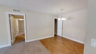 This single-floor space presents a straightforward and functional layout finished with a warm, neutral color palette throughout. The interior features a combination of natural hardwood flooring and plush carpeting, creating a clear distinction between living zones while maintaining a cohesive aesthetic. Walls are painted in a soft beige tone that enhances the sense of openness and pairs naturally with the warm wood tones underfoot.

A elegant multi-bulb pendant chandelier with brushed nickel finishes serves as a focal point in the dining or main area, providing both ambient and task lighting. The textured popcorn ceiling adds a classic touch characteristic of the architectural period.

An open doorway leads to an adjacent carpeted room, while a second doorway reveals a walk-in closet with visible shelving, offering practical storage solutions. White trim and baseboards frame each room cleanly, adding a crisp contrast to the neutral walls.

The overall ambiance is calm and move-in ready, with a clean, uncluttered environment that allows prospective occupants to envision their own furnishings and personal style. The space is well-suited for comfortable everyday living.