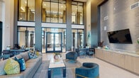 This contemporary amenity space makes an immediate impression with its dramatic double-height ceilings and expansive floor-to-ceiling glass walls that flood the interior with natural light. The grand lobby serves as the heart of the space, anchored by a sophisticated seating arrangement featuring plush gray sofas and teal accent chairs with gold-toned legs, centered around a sleek coffee table atop a textured area rug. A striking feature wall clad in layered tile houses a mounted television and a linear fireplace, while vibrant teal abstract artwork adds color throughout. Wide-plank light wood flooring and large-format tile flow seamlessly through the space, complementing the cool gray and neutral palette. A dedicated reception desk provides a polished point of arrival near the glass-framed entry doors. A secondary lounge area showcases additional seating, curated wall art, and a multi-screen media wall with a fireplace below, bathed in sunlight through soaring windows. The fully equipped catering kitchen features light wood cabinetry, stone countertops, stainless steel appliances, and a textured tile accent wall with integrated lighting. Glass-enclosed rooms, including a private conference room with a large meeting table and sheer-draped windows, offer functional, professional spaces within the broader communal environment.