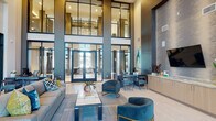 This contemporary amenity space makes an immediate impression with its dramatic double-height ceilings and expansive floor-to-ceiling glass walls that flood the interior with natural light. The grand lobby serves as the heart of the space, anchored by a sophisticated seating arrangement featuring plush gray sofas and teal accent chairs with gold-toned legs, centered around a sleek coffee table atop a textured area rug. A striking feature wall clad in layered tile houses a mounted television and a linear fireplace, while vibrant teal abstract artwork adds color throughout. Wide-plank light wood flooring and large-format tile flow seamlessly through the space, complementing the cool gray and neutral palette. A dedicated reception desk provides a polished point of arrival near the glass-framed entry doors. A secondary lounge area showcases additional seating, curated wall art, and a multi-screen media wall with a fireplace below, bathed in sunlight through soaring windows. The fully equipped catering kitchen features light wood cabinetry, stone countertops, stainless steel appliances, and a textured tile accent wall with integrated lighting. Glass-enclosed rooms, including a private conference room with a large meeting table and sheer-draped windows, offer functional, professional spaces within the broader communal environment.