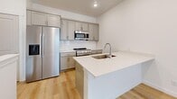 Located on W Wrigley Drive in Lehi, Utah, this well-designed multi-level home offers one bedroom and two bathrooms across three floors, making smart use of every square foot. The ground level provides an attached garage with automatic opener, drywall-finished walls, and direct interior access — offering secure parking and additional storage capacity.

The main living floor hosts an open-concept dining room, kitchen, and living area centered around a sleek, modern kitchen. The kitchen features gray shaker-style cabinetry, white quartz countertops, a kitchen island with an undermount sink, white subway tile backsplash, and stainless steel appliances including a gas range, microwave, and refrigerator. Light hardwood-style flooring ties the space together and enhances the bright, airy atmosphere.

The upper floor is home to the spacious primary bedroom, finished with plush neutral carpet, recessed lighting, and a window that brings in natural light. Two closets provide generous storage. The dedicated laundry room on this level includes side-by-side full-size washer and dryer units with overhead shelving and tile flooring. Both bathrooms are finished in clean, neutral tones — one featuring a soaking tub with built-in shelving niches and chrome fixtures. A balcony adds an inviting outdoor retreat to complete this efficiently designed home.