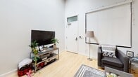 Located at 1030 Fifth Ave in Pittsburgh, Pennsylvania, this single-floor unit showcases a distinctive loft-style design characterized by soaring ceilings with exposed wooden beams, crisp white walls, and light hardwood flooring throughout. The open layout creates a generous sense of space, connecting the main living and kitchen areas seamlessly.

The living area is bright and functional, featuring high ceilings that enhance the airy atmosphere. A projection screen mounted on the wall serves as a standout entertainment feature, complemented by ample room for seating arrangements. The kitchen is well-equipped with rich warm-toned wood cabinetry, stainless steel appliances including a full-size refrigerator, electric range, and microwave, along with a sink and generous counter space—all set against the same light hardwood flooring.

The bedroom continues the loft aesthetic, with exposed beam ceilings, a window allowing natural light, a closet, and enough space for a full sleeping and workspace setup. Two bathrooms provide practical utility; one features a wide vanity with a stone-composite countertop and large mirror, while the other offers a shower/tub combination. Both include tile flooring. The overall ambiance is modern, urban, and versatile.