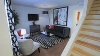 This two-story space offers a thoughtfully designed interior that balances contemporary style with functional living. The main level features a living area finished with rich hardwood flooring that anchors the room with warmth and contrast against the light-toned walls. A tufted charcoal sofa serves as the focal point of the seating arrangement, paired with a patterned accent chair and a bold black-and-white chevron area rug that adds graphic energy to the space. A large-scale abstract artwork with dramatic brushstrokes of black, red, and white commands attention on the main wall, while a wall-mounted flat-screen television and a dark storage dresser provide both entertainment and functional storage. A sleek arc floor lamp adds ambient lighting, complementing the overall modern aesthetic. A glass coffee table completes the living arrangement, adding an airy, open quality to the seating area. A staircase with light wood treads is visible along one edge of the room, providing access to the second floor. The overall ambiance is bold and curated, with a strong interplay of dark and light tones throughout.