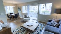 This single-floor residence offers a thoughtfully designed open-plan layout that seamlessly connects the living and dining areas, creating a cohesive and inviting atmosphere. The living area is anchored by hardwood-style flooring and features a geometric-patterned area rug in muted tones of blue, green, and gold, complementing the neutral color palette throughout. A substantial square wood coffee table serves as the centerpiece, paired with a plush sofa and a clean-lined accent chair in crisp white upholstery.

Natural light floods the space through large windows and a full-width sliding glass door, which opens to a patio overlooking a well-maintained landscaped courtyard. The abundant glazing creates a strong visual connection to the outdoors, enhancing the sense of openness and airiness. The dining area, positioned adjacent to the living space, accommodates a round table with dark accent chairs, ideal for everyday dining or entertaining. Soft, warm wall tones and minimal ornamentation give the interior a contemporary yet welcoming feel. Pendant lighting adds a stylish finishing touch. Overall, this space balances comfort, functionality, and modern design in a bright, light-filled setting.