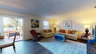 This well-appointed single-floor residence offers a thoughtfully laid-out interior with a warm, coastal-inspired aesthetic. The living room serves as a welcoming centerpiece, featuring wood-look flooring, wainscoting, multiple windows that invite natural light, and comfortable seating arranged around a distinctive trunk-style coffee table. Tropical artwork and colorful accent pillows lend the space a relaxed, inviting character.

The dining area flows naturally from the living space and is highlighted by sliding glass doors that open to an outdoor patio, offering pleasant views and easy indoor-outdoor access. Adjacent to the dining area, the kitchen is efficiently organized with ample white cabinetry, full appliances including a refrigerator, range, microwave, and dishwasher, along with a pass-through window and wood-look flooring.

The primary bedroom is generously sized with plush carpeting, a bold red dresser, and direct access to an en-suite bathroom and a walk-in closet. A second bedroom accommodates twin beds and features built-in closet storage, making it ideal for guests. Two full bathrooms, each with a tub-shower combination, serve the home. The overall ambiance is bright, cohesive, and comfortably furnished throughout.