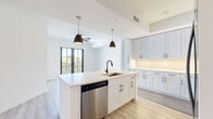 Located along Tamiami Trail in Naples, Florida, this single-floor unit showcases a clean, contemporary design characterized by crisp white walls, light wood-style flooring, and a cohesive modern aesthetic throughout. The open-concept layout connects the kitchen and living areas seamlessly, creating a bright and airy atmosphere enhanced by natural light from black-framed windows and glass doors.

The kitchen is a standout feature, equipped with white shaker-style cabinetry extending floor to ceiling, under-cabinet lighting, light quartz countertops, and matte black hardware. A central island with a built-in sink and stainless steel dishwasher provides additional workspace and storage. Stainless steel appliances, including a range, microwave, and French door refrigerator, complete the well-appointed space.

The living area opens directly to a private balcony through striking black-framed French doors, offering outdoor access and natural ventilation via a ceiling fan overhead. The bedrooms are generously sized with large windows, ceiling fans, and ample wall space. The bathroom features a dual-sink vanity with quartz countertops, a large framed mirror, and white cabinetry with black hardware. Throughout the unit, the consistent use of light tones and modern finishes creates a polished, move-in-ready environment.