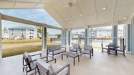 Located in Summerville, South Carolina, this single-story outdoor pavilion serves as a central community gathering space designed for relaxation and socializing. The structure features a striking vaulted gable roof with a clean white coffered ceiling, recessed lighting, and a ceiling fan that ensures comfort in the warm South Carolina climate. Distinctive blue-and-white board-and-batten exterior panels give the pavilion a fresh coastal aesthetic that complements the surrounding residential buildings. The open-air design is defined by wide white columns with blue accent panels, allowing unobstructed views of the pool area and the lush community grounds beyond. A built-in wet bar with a stone countertop and sink is positioned along the back wall beneath a wall-mounted flat-screen TV, making the space ideal for entertaining. Comfortable cushioned lounge seating and coffee tables are arranged throughout, creating inviting conversation areas. Additional dining-height seating provides flexible use of the space. Beyond the pavilion, the community grounds feature manicured open lawns, a decorative fountain pond, and mature trees, all framed by multi-story residential buildings in a coordinating coastal color palette. The poolside location, combined with the pavilion's thoughtful amenities and open design, make it a functional and welcoming outdoor retreat.