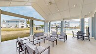 Located in Summerville, South Carolina, this single-story outdoor pavilion serves as a central community gathering space designed for relaxation and socializing. The structure features a striking vaulted gable roof with a clean white coffered ceiling, recessed lighting, and a ceiling fan that ensures comfort in the warm South Carolina climate. Distinctive blue-and-white board-and-batten exterior panels give the pavilion a fresh coastal aesthetic that complements the surrounding residential buildings. The open-air design is defined by wide white columns with blue accent panels, allowing unobstructed views of the pool area and the lush community grounds beyond. A built-in wet bar with a stone countertop and sink is positioned along the back wall beneath a wall-mounted flat-screen TV, making the space ideal for entertaining. Comfortable cushioned lounge seating and coffee tables are arranged throughout, creating inviting conversation areas. Additional dining-height seating provides flexible use of the space. Beyond the pavilion, the community grounds feature manicured open lawns, a decorative fountain pond, and mature trees, all framed by multi-story residential buildings in a coordinating coastal color palette. The poolside location, combined with the pavilion's thoughtful amenities and open design, make it a functional and welcoming outdoor retreat.