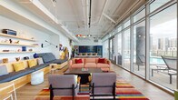 This contemporary single-floor amenity space is thoughtfully designed to blend energetic style with functional comfort. The open-plan layout is anchored by expansive floor-to-ceiling glass walls along one side, flooding the interior with natural light and offering sweeping views of the surrounding cityscape and an outdoor terrace complete with lounge seating and a pool.

The main lounge area is a visual centerpiece, featuring a large-scale, colorful mural that enlivens the white walls, complemented by a bold striped area rug in warm, vivid tones. Seating arrangements include a salmon-toned sofa with red accent pillows, a built-in banquette with curated display shelving, and additional armchairs grouped around a dark coffee table — offering versatile social and relaxation zones.

A secondary seating nook near the glass facade features an oversized navy sectional sofa with rust-colored accent pillows, positioned to take full advantage of the panoramic outdoor views. The entrance area is welcoming, with a striking geometric black glass door and intimate seating.

The kitchen area is fully equipped with Bosch double wall ovens, a deep blue glossy tile backsplash, open wooden floating shelves, and warm wood cabinetry with grey countertops. Exposed ceiling infrastructure with white-painted ductwork and pendant lighting adds an industrial-chic character throughout the space. Warm wood-look flooring unifies the entire layout cohesively.