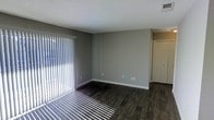 This single-floor residence presents a straightforward and well-maintained interior with a clean, neutral aesthetic throughout. The main living area is finished with rich, dark hardwood flooring that contrasts crisply against the light gray walls and white baseboards, creating a modern and cohesive look. A textured popcorn ceiling adds a classic touch overhead, while a ceiling-mounted HVAC vent ensures year-round climate comfort. Natural light floods the space through a large sliding glass door dressed with vertical blinds, casting bright, defined light patterns across the floor and lending the room an open, airy feel. The layout is open and unobstructed, allowing for flexible furniture arrangements to suit a variety of needs. A closet with bifold doors is visible adjacent to the main living area, providing convenient built-in storage. The neutral color palette of soft grays and whites throughout creates a calm, versatile backdrop that accommodates a wide range of interior styles. Overall, the space is practical, well-lit, and move-in ready, offering a comfortable and adaptable living environment.