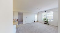 This single-floor residence is designed with a clean, straightforward layout that maximizes functionality throughout. The living area is generously proportioned, featuring neutral-toned wall-to-wall carpeting, white walls, and multiple windows that allow natural light to fill the space. A glass-panel door provides additional brightness and connects the interior to the outside. The open layout transitions seamlessly to the kitchen, which is visible from the living area via a peninsula counter with a light-colored laminate surface. The kitchen is equipped with warm oak cabinetry offering ample upper and lower storage, white appliances including an electric range, dishwasher, and refrigerator, complemented by light tile flooring and overhead fluorescent lighting. The bedrooms are carpeted and maintain the home's neutral palette, each offering generous wall space and natural light from windows with blinds. One bedroom features an open closet with a built-in shelving rod. The bathroom is finished with white tile flooring, a single-sink oak vanity with a white countertop, a framed mirror, and a full-size tub and shower combination. The overall ambiance is bright, clean, and move-in ready.