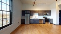This single-floor loft-style unit showcases an open, airy layout with standout industrial-chic architecture. Soaring ceilings with a white-painted wood plank finish and exposed red fire suppression pipes add a distinctive character throughout the space. Warm-toned hardwood flooring runs continuously, grounding the open floor plan with natural texture and cohesion.

The kitchen is integrated seamlessly into the main living area and is outfitted with a full suite of stainless steel appliances, including a refrigerator, electric range, built-in microwave, and dishwasher. Dark espresso-toned cabinetry extends both above and below the counters, complemented by clean white countertops and a stainless steel sink. Track lighting positioned above provides excellent task illumination for the kitchen zone.

Large, multi-pane black-framed windows in the main living area invite generous natural light and offer views of the surrounding landscape, enhancing the sense of openness. The bedroom is a generously proportioned, private space that features high ceilings echoing the main living area, a wall-mounted split HVAC unit for individual climate control, and direct access to a walk-in closet. The bathroom includes dark cabinetry with a white countertop vanity and a spacious adjacent walk-in closet with built-in shelving and hanging storage. The overall ambiance is modern, clean, and functional with an industrial edge.