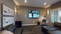 Located at 5927 Avery Wood Ave in Dublin, Ohio, this single-floor space is designed as a dedicated media and entertainment room with a polished, cinema-inspired aesthetic. The screening area is anchored by a large wall-mounted flat-screen TV flanked by in-wall speakers, creating an immersive audio-visual setup ideal for movie nights and gatherings. Warm sconce lighting lines the neutral gray walls, casting an inviting ambiance that complements the recessed ceiling lights above. The room is furnished with generously sized dark leather sectional sofas and ottomans, offering comfortable seating for multiple guests, all arranged atop a richly patterned carpet. A classic popcorn machine on a vintage-style cart adds a charming, theater-inspired touch to the space. An adjacent area, visible through a wide doorway, features light wood-look flooring, striped accent chairs, and abundant natural light streaming through large windows dressed with elegant drip curtains and blinds, providing a bright contrast to the darker media room. Framed artwork adorns the walls, adding personality and character throughout. The thoughtful layout and cohesive design make this space a well-appointed venue for entertainment and relaxation.