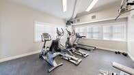 This fitness center in Evans, Colorado offers a well-appointed single-room exercise space designed to support a comprehensive workout routine. The room features soaring ceilings that lend an open, airy feel despite its compact footprint, with neutral-toned walls and durable rubber flooring suited for high-traffic athletic use.

Natural light flows in through multiple large windows fitted with white horizontal blinds, creating a bright and energizing environment. A wall-mounted flat-screen television provides entertainment during cardio sessions, while a large mirror along one wall visually expands the space and allows users to monitor their form.

The cardio section is equipped with a Matrix treadmill, elliptical trainers, and a recumbent stationary bike, offering a variety of low-impact and high-intensity options. A multi-station cable and weight machine anchors the strength training area, complemented by free weights including a barbell and additional accessories for targeted resistance training.

Overhead fluorescent lighting ensures consistent illumination throughout, and the room is climate-controlled with visible ventilation units. A private entry door with an adjacent window provides convenient and secure access. Overall, this fitness center delivers a functional and comfortable environment for residents seeking on-site exercise amenities in Evans, CO.