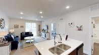 This single-floor space showcases an open-concept layout that connects the kitchen, dining, and living areas into one cohesive, well-lit environment. The kitchen features a large island with a double-basin stainless steel sink, light quartz countertops, and dark cabinetry, offering both functional workspace and a natural gathering point. Recessed lighting throughout the ceiling provides consistent, even illumination that complements the abundant natural light streaming in through large sliding glass doors in the living area, which open to an outdoor space beyond.

The living room is generously proportioned, with neutral carpet, a cozy seating arrangement anchored by a navy sofa and round white coffee table, and a farmhouse-style media console that adds a touch of rustic charm. A bedroom is visible through an interior doorway, suggesting a practical and well-organized floor plan. The walls are painted in a clean, soft white that enhances the sense of openness throughout. Overall, the space presents a modern, move-in-ready aesthetic with a warm, inviting ambiance suited for comfortable everyday living.