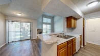 This two-story residence offers 2 bedrooms and 2 bathrooms across a well-organized layout spanning two floors. The main level encompasses the primary living areas, including an open-concept kitchen and living room that together form the heart of the home. The living room stands out with dramatic vaulted ceilings, large windows that invite abundant natural light, a sliding glass door leading to an outdoor area, and a striking stone-surround fireplace that serves as a focal point. The kitchen features warm wood cabinetry, speckled stone countertops, a peninsula with a built-in sink, and white appliances, with ample cabinet storage lining both sides of the galley-style workspace. A carpeted staircase connects the main level to the upper floor, where both bedrooms are located. The primary bedroom is a comfortable, well-proportioned room with plush carpet, a closet, and natural light from a window. The second bedroom similarly features carpet flooring and closet access. Both bathrooms are functional and clean, with one featuring a double-sink vanity with stone countertops and mirrors, and the other offering a full tub-shower combination. Hardwood-style flooring runs throughout the main level, complementing the home's neutral color palette and textured ceilings.