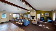 This single-floor community clubhouse at The Ponds at Georgetown is designed to foster comfort and social interaction through a thoughtfully arranged, open-concept layout. The main lounge is the centerpiece of the space, showcasing dramatic vaulted ceilings accented by bold dark wood beams, track lighting, and a modern chandelier that together create a bright and airy ambiance. A striking floor-to-ceiling stacked stone fireplace with a wood mantel serves as the focal point of the room, flanked by large windows dressed in gray drape curtains that allow abundant natural light to fill the space. Olive green accent walls add a vibrant, contemporary energy, while rich patterned carpeting grounds the seating arrangement of blue sofas, neutral accent chairs, and glass-top coffee tables. A dedicated dining area near the windows offers round tables and eclectic patterned chairs, ideal for casual gatherings. Adjacent to the main lounge, a well-appointed kitchenette features dark espresso cabinetry, granite countertops, stainless steel appliances including a microwave, range, and dishwasher, along with a coffee station — providing all the essentials for hosting events and welcoming residents. Hardwood-style flooring connects the kitchenette to the lounge, completing this polished and functional community space.