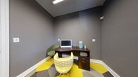 This single-floor office space is thoughtfully designed to maximize functionality within a compact footprint. The room is defined by its striking flooring — a bold geometric pattern of yellow and gray hexagonal carpet tiles that energizes the space and adds a modern design flair. The walls are finished in a deep charcoal gray, creating a focused, professional atmosphere complemented by crisp white baseboards and trim throughout.

The primary workstation is a dark walnut-toned corner desk, providing ample surface area for a desktop computer setup, along with integrated drawers and open shelving for organized storage. Two accent chairs — one in sage green and one in a yellow geometric print — provide comfortable seating options that tie the color palette together.

A window fitted with horizontal wood-toned blinds allows for natural light and a view of greenery outside, adding a refreshing element to the workspace. An additional built-in credenza along one wall features light wood cabinetry, granite-style countertops, and a decorative tile backsplash, offering extra storage and surface space.

An adjacent tiled area is visible through the doorway, suggesting a broader facility context. Overall, this office space balances practical workstation design with vibrant, carefully chosen décor elements.