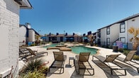 Located at 9669 Forest Ln in Dallas, Texas, this outdoor amenity area serves as a central gathering space within a multi-building residential community. The centerpiece is a generously sized freeform swimming pool with a natural stone-edged spa and a decorative fountain feature, creating a resort-like atmosphere under the open Texas sky. The pool's organic shape and clear blue-green water are complemented by a spacious concrete sun deck that surrounds the area, offering ample room for relaxation and recreation. Numerous lounge chairs and small side tables are arranged throughout the deck, providing comfortable spaces for sunbathing and leisure. Vibrant blue market umbrellas are stationed around the perimeter, offering shaded seating areas for those seeking relief from the sun. The community buildings that frame the space feature a mix of white stucco and blue siding exteriors, pitched rooflines, and multiple chimneys, contributing to a cohesive architectural aesthetic. Mature palm trees and drought-tolerant landscaping, including ornamental grasses and yucca plants, line the walkways and fenced boundaries, adding a layer of greenery and texture. Black iron fencing encloses the area, ensuring security while maintaining open sightlines throughout this well-appointed outdoor retreat.