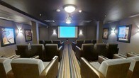 This private screening room is designed to deliver a true cinematic experience in an intimate, well-appointed setting. The room is outfitted with rows of generously sized, upholstered theater-style chairs arranged in a forward-facing layout, providing comfortable and unobstructed sightlines to the large front projection screen. The ceiling-mounted projector anchors the room's purpose and capability, while flush ceiling light fixtures and wall-mounted sconces provide ambient lighting that can be adjusted to suit any screening environment. The dark, neutral tones of the walls and ceiling create a focused atmosphere that minimizes distraction and enhances on-screen contrast. A bold striped carpet runner runs the length of the central aisle, adding a visual accent that complements the rich brown and tan palette of the seating. Framed artwork on the side walls adds a polished, curated touch to the space. The room's single-floor layout ensures accessibility, and the overall design balances functionality with comfort, making it well-suited for movie screenings, presentations, or private events requiring an engaging visual display.