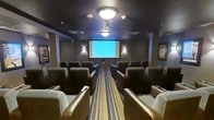 This private screening room is designed to deliver a true cinematic experience in an intimate, well-appointed setting. The room is outfitted with rows of generously sized, upholstered theater-style chairs arranged in a forward-facing layout, providing comfortable and unobstructed sightlines to the large front projection screen. The ceiling-mounted projector anchors the room's purpose and capability, while flush ceiling light fixtures and wall-mounted sconces provide ambient lighting that can be adjusted to suit any screening environment. The dark, neutral tones of the walls and ceiling create a focused atmosphere that minimizes distraction and enhances on-screen contrast. A bold striped carpet runner runs the length of the central aisle, adding a visual accent that complements the rich brown and tan palette of the seating. Framed artwork on the side walls adds a polished, curated touch to the space. The room's single-floor layout ensures accessibility, and the overall design balances functionality with comfort, making it well-suited for movie screenings, presentations, or private events requiring an engaging visual display.