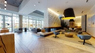 This contemporary single-floor lobby and amenity lounge is designed with a refined yet welcoming aesthetic. The space is anchored by a striking textured white sculptural wall surrounding a sleek double-sided fireplace, creating a dramatic focal point that divides the open-plan interior into distinct seating zones. Chevron-patterned tile flooring runs throughout, adding visual depth and continuity across the space.

Natural light pours in through expansive floor-to-ceiling glass walls and large sliding doors that open directly to an outdoor courtyard and pool area, offering seamless indoor-outdoor connectivity. The lounge is furnished with modern upholstered chairs, a classic leather lounge chair, round live-edge wood coffee tables, and stylish area rugs — all thoughtfully arranged to encourage both social interaction and relaxed individual use.

Oversized pendant light fixtures with bold yellow interiors add a pop of color and warmth to the seating areas. Curated artwork decorates accent walls throughout. A separate bar area features a thick reclaimed wood countertop, leather-topped barstools, pendant lighting, and dark tile backsplash against deep navy walls, creating an intimate, lounge-like atmosphere. Entryways are framed by glass-paneled double doors, concrete columns, and rich wood panel accents, completing the space's modern industrial character.
