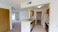 This single-floor residence is designed with an open and functional layout that seamlessly connects the kitchen and living areas. The kitchen is a standout feature, equipped with warm wood cabinetry that provides ample storage, a peninsula-style countertop with a double basin sink, and a dishwasher beneath the counter. A full-size refrigerator anchors one end of the kitchen, while tile flooring underfoot adds durability and ease of maintenance. The peninsula counter creates a natural division between the kitchen and the adjoining living space, making the layout ideal for everyday living. A solid wood entry door with a lower ventilation panel adds a classic, sturdy element to the space. The living area transitions smoothly from the kitchen, featuring plush carpeting that adds warmth and comfort. A sliding glass door floods the living space with natural light and offers a view to the outdoors. Overhead, flush-mount lighting and centrally placed HVAC vents ensure consistent comfort throughout. The clean white walls and neutral tones throughout the space create a bright, welcoming ambiance that is both versatile and easy to personalize.