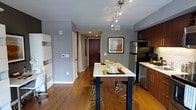 This single-floor space is thoughtfully designed to maximize functionality within a compact footprint. The open-plan living and kitchen area serves as the heart of the unit, anchored by a sleek white-topped dining table with black metal legs, accompanied by matching bar-height stools — ideal for casual dining or entertaining.

The kitchen is outfitted with dark wood cabinetry, stainless steel appliances including a full-sized refrigerator and dishwasher, and a clean white countertop workspace. A track lighting fixture above the dining area provides focused illumination, complementing the recessed ceiling lights throughout.

Warm hardwood flooring runs throughout the main space, adding richness against the cool grey-painted walls. A hallway leads toward the entry door — a rich dark wood with a keypad lock — flanked by additional doorways suggesting bedroom and bathroom access. A framed artwork piece and subtle decorative accents add a touch of personality. The overall ambiance is modern, clean, and efficiently arranged for comfortable everyday living.