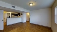 This single-floor residence offers a well-designed, open-concept layout that maximizes both space and functionality. The main living area is anchored by warm hardwood flooring that extends throughout, creating a cohesive and inviting atmosphere. Crisp white walls and flush ceiling lighting contribute to a bright, clean aesthetic that feels open and airy.

A defining feature of the layout is the kitchen pass-through, which connects the cooking area to the main living space, promoting an effortless flow between rooms. The kitchen is equipped with dark cabinetry that provides a rich contrast against the lighter surroundings, along with under-cabinet lighting that adds a warm glow to the workspace. A visible countertop peninsula further enhances the connection between the kitchen and living area, making it ideal for casual dining or entertaining.

An arched doorway leads from the living space into a hallway, hinting at additional rooms beyond and adding a subtle architectural charm to the interior. HVAC vents are integrated into the ceiling, ensuring year-round climate comfort. The overall design of the space balances practicality with a welcoming ambiance, making it a comfortable and functional environment suited for everyday living.