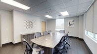 This single-floor meeting space is designed to support professional collaboration in a clean, contemporary setting. At its center sits a generously sized, wood-toned boardroom table surrounded by ergonomic task chairs, comfortably accommodating up to eight participants. The room is finished with dark tile flooring and neutral grey walls, creating a polished yet understated aesthetic.

Recessed panel lighting in the drop ceiling provides consistent, even illumination throughout the space, complemented by natural light streaming through roller-shaded windows that offer views of the surrounding exterior. A wall-mounted flat-screen display at one end of the room supports presentations and video conferencing needs.

Decorative wall art featuring abstract brushstroke compositions adds a subtle creative touch, while a low-profile storage credenza along one wall provides practical utility. A warm-toned wood door offers a private entry point, reinforcing the room's sense of enclosure and focus. Overall, the space strikes a balance between functional design and professional ambiance, making it well-suited for meetings, presentations, or collaborative work sessions.