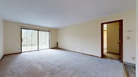 Located at 4514 Farringdon Rd NW in Canton, Ohio, this single-floor property offers a straightforward, functional layout with a clean and neutral interior palette. The living area is generously proportioned, featuring plush wall-to-wall carpeting and warm cream-toned walls that contribute to a bright, open atmosphere. A full-width sliding glass door fitted with vertical blinds allows natural light to fill the space while providing outdoor access and views of surrounding greenery.

The transition between the living area and adjacent spaces is defined by a shift in flooring, moving from carpet to updated light wood-look luxury vinyl plank, which extends through the hallways and into the kitchen. Rich, warm-toned wood doors with brushed nickel hardware provide visual contrast against the neutral walls throughout the unit.

The galley-style kitchen is efficiently organized, featuring medium-toned wood cabinetry with ample upper and lower storage, white countertops, a stainless sink, and a full suite of appliances including a dishwasher, range, and refrigerator. A dropped ceiling with panel lighting completes the kitchen's functional design. The overall layout of the space is practical and well-connected, offering a cohesive flow between the primary living areas.