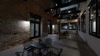 Located at 698 Martin Luther King Jr Blvd in the heart of Macon, Georgia, this distinctive space offers a compelling blend of historic industrial character and atmospheric design. The hallway functions as an open-air courtyard corridor, defined by original exposed brick walls that rise to impressive heights, lending the space a raw, authentic charm rooted in its architectural heritage.

Overhead, bold black steel beams span the upper levels, complemented by string globe lights that drape across the space to create a warm, ambient glow. An upper-level balcony railing is visible above, hinting at the multi-story nature of the surrounding structure. Large black-framed windows and glass panel walls allow views into adjacent interior spaces, bridging the connection between indoors and out.

The floor is finished in stone-toned tile throughout, providing a clean, cohesive foundation that contrasts beautifully with the textured brick. The courtyard is furnished with multiple seating arrangements — including wicker and metal bistro-style tables and chairs — making it well-suited for social gatherings or casual use. A sliding barn door adds a modern-rustic accent, while wall-mounted sconces provide additional focused lighting. The overall ambiance is moody, intimate, and rich with historic character.