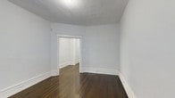 This single-floor space presents a clean, understated aesthetic defined by its classic architectural elements and well-maintained finishes. The interior features rich, dark hardwood floors that run throughout, providing warmth and character against the crisp white walls and ceilings. Generous white baseboards add a traditional touch, complementing the simple yet elegant door trim that frames the passageway connecting the rooms. The open doorway leads to an adjoining room, suggesting a flowing, connected layout that allows for versatile use of the space. The smooth white ceilings, lit by a ceiling-mounted light fixture, contribute to an airy, well-lit atmosphere. The walls are painted in a soft, neutral light gray-white tone, creating a bright and calming ambiance throughout. The baseboard heating unit along the wall indicates year-round comfort. The space is notably open and uncluttered, offering a blank canvas that can be adapted to a variety of uses. Overall, the combination of hardwood flooring, classic trim work, and neutral palette gives this space a timeless quality with strong functional potential.