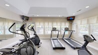 Located at 4901 Warner Rd in Columbus, Ohio, this single-floor fitness center is thoughtfully designed to accommodate a comprehensive range of workout routines. The space is organized into distinct exercise zones, providing ample room for both cardio and strength training activities. The cardio area features multiple treadmills, elliptical machines, and stationary bikes, positioned along a wall of windows dressed with Roman shades that invite generous natural light and offer views of the surrounding outdoor area. Wall-mounted televisions are positioned for easy viewing during workouts. The strength training section is equipped with a multi-station cable machine, a weight bench, and additional resistance equipment, all set against a large wall mirror that enhances the sense of openness throughout the space. Recessed lighting throughout the vaulted ceiling keeps the room bright and well-lit at all times. Neutral-toned walls and plush carpeted flooring contribute to a clean, welcoming atmosphere. A water fountain is conveniently located within the space, along with restroom access. The overall layout is practical and efficient, making this fitness center a well-appointed amenity for those seeking a consistent and comfortable exercise environment.