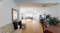 This expansive single-story space is designed to accommodate a variety of activities within an open, well-connected layout. The interior is unified by warm hardwood flooring throughout and soft, light-gray walls that contribute to a bright and welcoming atmosphere.

The main gathering area is anchored by a full-size pool table positioned beneath soaring vaulted ceilings, which are fitted with recessed lighting and a ceiling fan. Multiple windows along the perimeter flood the space with natural light and offer views of surrounding greenery. A comfortable lounge area with seating and a workstation featuring ladder shelving and a desk provide functional zones within the open floor plan.

A dedicated dining area includes a large wooden table with cross-back chairs, set within a bay-window alcove that further enhances natural illumination. Adjacent to this is a well-equipped kitchen featuring dark cabinetry, light countertops, stainless steel appliances, and a pass-through window that maintains visual connectivity with the main space.

Overall, the space balances recreational and functional use effectively, with a clean, airy design aesthetic supported by vaulted architecture, ample windows, and versatile furnishing arrangements.