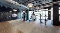 This contemporary fitness center is thoughtfully designed to accommodate a wide range of workout preferences within a single, expansive floor. The space is defined by an industrial-chic aesthetic, with exposed ductwork and piping set against high ceilings, complemented by large pendant light fixtures that provide warm, even illumination throughout.

The main workout floor is divided into distinct functional zones. A dedicated strength training area features a variety of resistance machines, a dumbbell rack, and open rubber-floored areas ideal for functional movements. TRX suspension trainers are mounted for bodyweight training, while a striking textured black tile accent wall adds a bold visual element and houses a mounted flat-screen display. Floor-to-ceiling windows along one wall flood the cardio zone with natural light and outdoor views, where treadmills and elliptical machines are positioned for an energizing workout experience.

Adjacent to the main floor, a separate studio space is enclosed with glass-panel walls and doors, housing a full arrangement of spinning bikes on light hardwood flooring. The studio is lined with wall-mounted mirrors and a ballet barre, making it versatile for both cycling classes and other group fitness activities. Dedicated restroom and changing facilities are accessible directly from the fitness floor, rounding out this comprehensive and professionally appointed fitness facility.