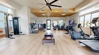 Located in Rancho Cucamonga, CA, this fitness center offers a thoughtfully designed, single-floor workout facility that combines modern aesthetics with a comprehensive range of exercise equipment. The space features light wood-look flooring throughout, creating a clean and inviting atmosphere that encourages activity.

A standout architectural element is the recessed wood-paneled ceiling section at the center of the room, accented by recessed lighting and a large ceiling fan that promotes airflow and comfort during workouts. The surrounding perimeter walls rise higher, fitted with large windows that allow abundant natural light to fill the room while offering views of the surrounding landscape, including palm trees and the broader Rancho Cucamonga skyline.

The fitness center is well-appointed with a diverse selection of equipment. Cardio machines include multiple treadmills, ellipticals, a rowing machine, and stationary bikes. The strength training area features a cable machine with a bench, a dumbbell rack with a full range of free weights, stability balls, and a flat utility bench. A large wall-mounted mirror expands the sense of space and allows users to monitor their form. A wall-mounted TV and a water fountain add further convenience. The main entrance features glass double doors, and the neutral, light-toned walls contribute to an open, energizing environment throughout.