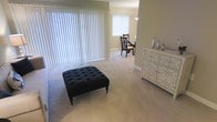 This single-floor residence offers a welcoming and functional layout designed for everyday living. The living area is finished with wall-to-wall neutral carpeting that adds warmth underfoot and contributes to the overall soft, comfortable atmosphere of the space. Large vertical blinds spanning a wide window wall allow for generous natural light while providing full privacy control, making the room feel bright and airy.

The open flow between the living and dining areas creates a sense of spaciousness, with the dining space anchored by a classic dark wood chair set and a pendant light overhead. Neutral-toned walls throughout maintain a clean and cohesive look that complements a variety of décor styles.

A decorative dresser with floral patterned drawer fronts adds a distinctive design touch, balancing the understated tones of the surrounding space. The tufted ottoman in the living area serves as a stylish focal point, offering both visual interest and practical function.

Overall, this residence presents a calm, livable environment with thoughtful design details. Its single-floor layout ensures ease of movement throughout, and the consistent use of neutral tones and soft textures gives the space a timeless, well-maintained character suited for comfortable daily living.