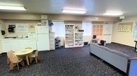 This single-floor community room is designed as a versatile, activity-oriented space that serves multiple functional purposes under one roof. The room features a compact kitchenette area equipped with a full-size refrigerator, microwave, and sink with under-counter cabinetry, providing practical utility for group activities and gatherings. Adjacent to the kitchenette is a children's activity zone, furnished with a small round table and child-sized chairs, ideal for structured learning or supervised play. A dedicated reading and resource area lines one wall, featuring well-stocked open bookshelves filled with books, educational materials, and supplies, alongside a multi-drawer filing cabinet and a rolling utility cart for organized storage. On the opposite end of the room, a lounge area offers comfortable seating with a sofa and a computer workstation, creating a space for relaxed interaction or independent work. The room is illuminated by recessed fluorescent ceiling fixtures and benefits from natural light through multiple windows fitted with horizontal blinds. The floor throughout is covered in patterned commercial-grade carpet, and the textured walls give the space a functional, community-center character. Overall, the layout is open and practical, supporting a range of educational, social, and recreational activities.