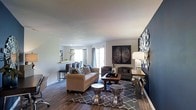 Located at 1390 Christopher Wren Dr in Gahanna, Ohio, this single-floor residence offers a well-appointed and thoughtfully designed living space. The main living area immediately draws attention with its rich hardwood flooring, which adds warmth and a sense of continuity throughout the room. Large windows allow generous amounts of natural light to pour in, creating a bright and inviting atmosphere, while sliding glass doors provide easy access to the outdoors. The living room is anchored by a bold accent wall in deep navy blue, offering a striking contrast against the neutral white and gray tones of the surrounding walls and ceiling. The open layout provides a comfortable sense of spaciousness, allowing for flexible furniture arrangements. A seamless transition connects the living area to a dining space visible in the background, enhancing the open-concept feel of the home. The overall ambiance is contemporary and refined, with a neutral color palette punctuated by decorative accents and layered textures. This space balances both style and functionality, making it a comfortable and visually engaging home in a well-established Gahanna neighborhood.