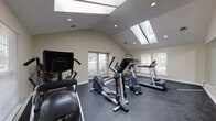 This dedicated fitness space is thoughtfully designed to support a complete workout experience within a single, open room. The standout architectural feature is the vaulted ceiling, which rises to an impressive height and is fitted with multiple skylights that flood the space with abundant natural light throughout the day. Recessed lighting complements the skylights, ensuring consistent illumination at all hours. The walls are finished in a neutral warm beige tone, contributing to a clean and focused atmosphere. Multiple windows along the perimeter walls provide additional natural light and outdoor views, enhancing the overall sense of openness. The floor is covered in durable rubber gym flooring with a speckled dark finish, offering both traction and impact absorption for high-intensity workouts. A large wall-mounted mirror spans a significant portion of one wall, visually expanding the room and allowing users to monitor their form during exercise. The space is equipped with a range of cardio and strength training machines, including treadmills, elliptical trainers, a stationary cycle, and a multi-function power tower for bodyweight exercises. The layout provides ample space between equipment, allowing for comfortable use of all machines simultaneously. A doorway connects the fitness room to adjacent areas of the building.
