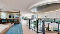 This expansive two-story amenity space is designed with a coastal-resort aesthetic, combining bold color, refined materials, and thoughtful architectural detailing throughout. The upper level is anchored by a striking oval mosaic ceiling installation with integrated ambient lighting, creating a dramatic focal point above the glass-paneled mezzanine railing that overlooks the lower level. Light wood-look flooring flows throughout the upper floor, complemented by vibrant teal-patterned carpeting that defines distinct lounge and activity zones.

The upper level features a well-appointed bar area with a mounted television, teal upholstered bar seating, and warm pendant lighting. A dedicated game room offers a full-size billiards table, wall-mounted TV, and ample open space, while a lounge zone with a sectional sofa and slatted wood room divider provides a relaxed retreat. A stylish communal dining area showcases a sculptural curved wood table, woven pendant lights, and views to the outdoors. A linear fireplace set into a floor-to-ceiling wood panel wall adds warmth near the entry.

The ground level includes a bright, tile-floored multipurpose room with floor-to-ceiling windows and a pool area visible from the mezzanine. Curated artwork, decorative glass wall pieces, and lush tropical greenery complete the polished, resort-inspired atmosphere.
