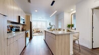 This contemporary single-floor residence is designed with a clean, modern aesthetic and an efficient, open layout. The heart of the home is a sleek, open-concept kitchen featuring light wood cabinetry, granite countertops, a generously sized center island with bar seating, and a full suite of stainless steel appliances. A linear pendant light runs above the island, complementing the recessed lighting overhead. Rich hardwood-style flooring flows seamlessly from the kitchen into the adjacent living area, creating a cohesive and inviting space. The living room is bright and airy, benefiting from multiple large windows fitted with white blinds that allow for generous natural light. The bedroom continues the light-filled theme, with a prominent window framing outdoor views and plush carpeting underfoot, offering a comfortable retreat. The bathroom is finished with wood-look flooring, a granite-topped vanity with light wood cabinetry matching the kitchen, and a tub-shower combination with white subway tile surround. Throughout the residence, the consistent use of neutral tones, natural textures, and modern fixtures creates a cohesive and welcoming atmosphere well-suited for comfortable everyday living.