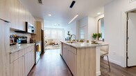 This contemporary single-floor residence is designed with a clean, modern aesthetic and an efficient, open layout. The heart of the home is a sleek, open-concept kitchen featuring light wood cabinetry, granite countertops, a generously sized center island with bar seating, and a full suite of stainless steel appliances. A linear pendant light runs above the island, complementing the recessed lighting overhead. Rich hardwood-style flooring flows seamlessly from the kitchen into the adjacent living area, creating a cohesive and inviting space. The living room is bright and airy, benefiting from multiple large windows fitted with white blinds that allow for generous natural light. The bedroom continues the light-filled theme, with a prominent window framing outdoor views and plush carpeting underfoot, offering a comfortable retreat. The bathroom is finished with wood-look flooring, a granite-topped vanity with light wood cabinetry matching the kitchen, and a tub-shower combination with white subway tile surround. Throughout the residence, the consistent use of neutral tones, natural textures, and modern fixtures creates a cohesive and welcoming atmosphere well-suited for comfortable everyday living.